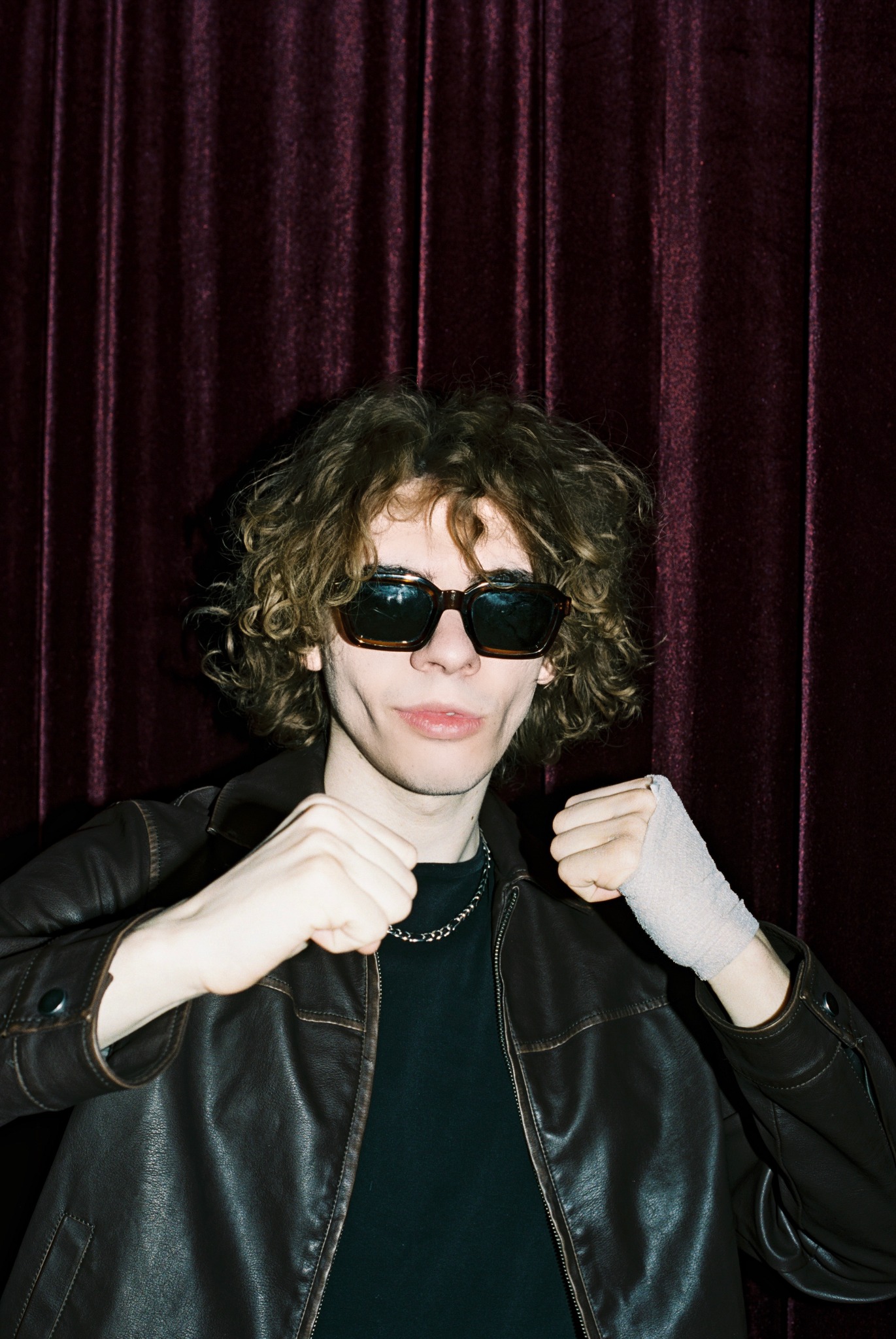 Caner Oz in sunglasses and a leather jacket against burgundy curtains
